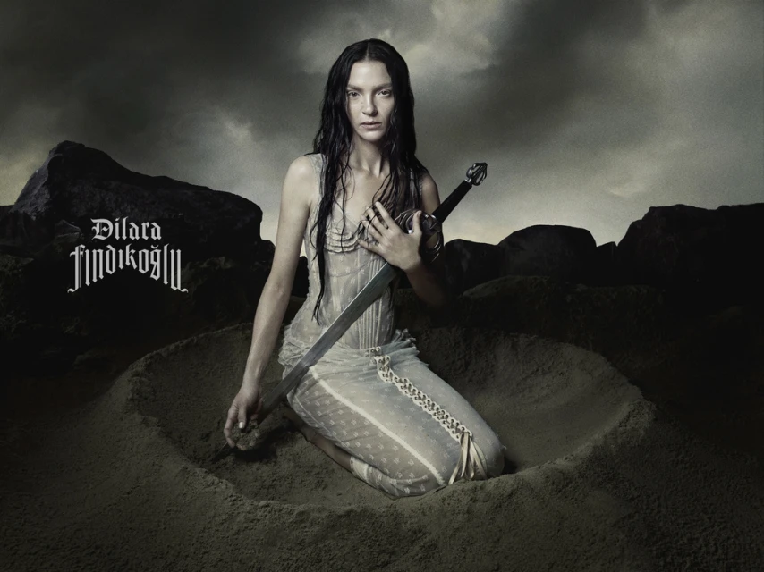 Dramatic portrait of a woman kneeling in sand, holding a sword, under a stormy sky. Dark, moody aesthetic with a gothic-inspired vibe.