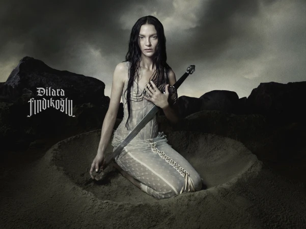 Dramatic portrait of a woman kneeling in sand, holding a sword, under a stormy sky. Dark, moody aesthetic with a gothic-inspired vibe.