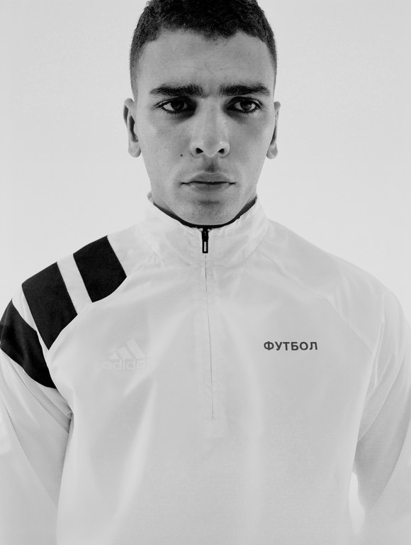 Close-up portrait of a young man in a white track jacket with black stripes against a white background in a studio setting.