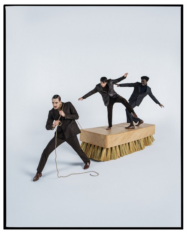Three men in suits pose with a large brush, one pulling with a rope, two balancing on top, in a studio setting with a light background.