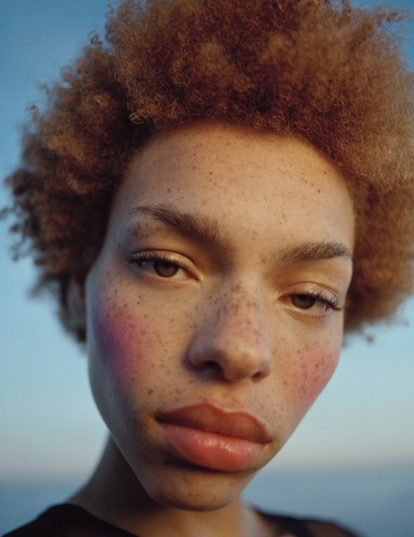 Close-up portrait of a young adult woman with freckles and curly auburn hair against a soft sky and ocean background, captured in natural light.