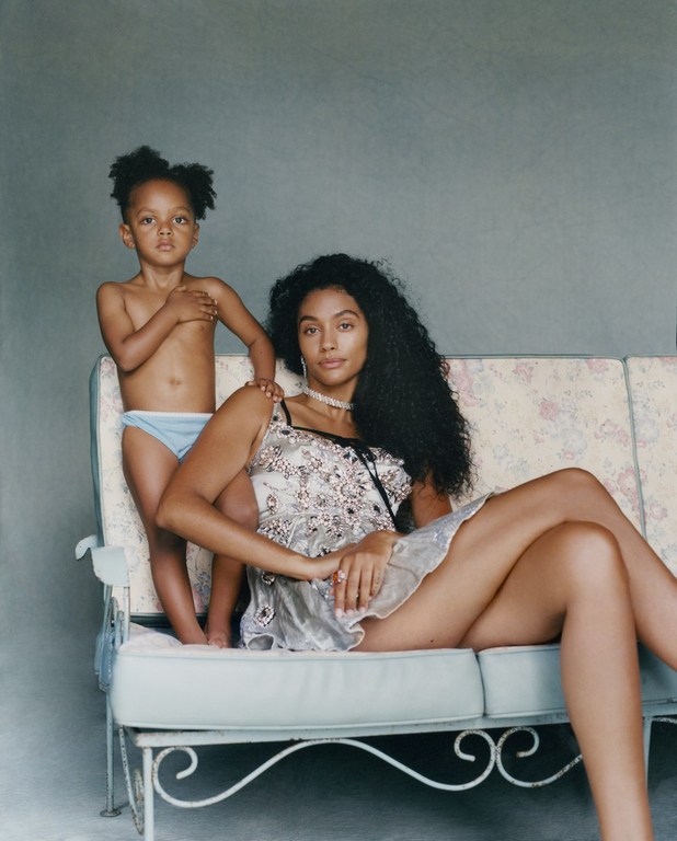 A mother and child pose on a vintage sofa in a studio, with the child standing behind the seated adult woman, both looking at the camera.