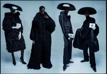Four Black models in avant-garde black outfits and oversized circular hats holding numbered placards against a grey backdrop