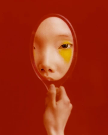Close-up portrait of a person holding an oval hand mirror reflecting their face with bright yellow eye makeup against a solid red background