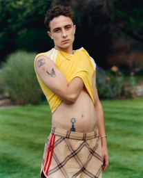 A young man with short dark hair wearing a yellow t-shirt and plaid shorts standing outdoors in a garden setting.