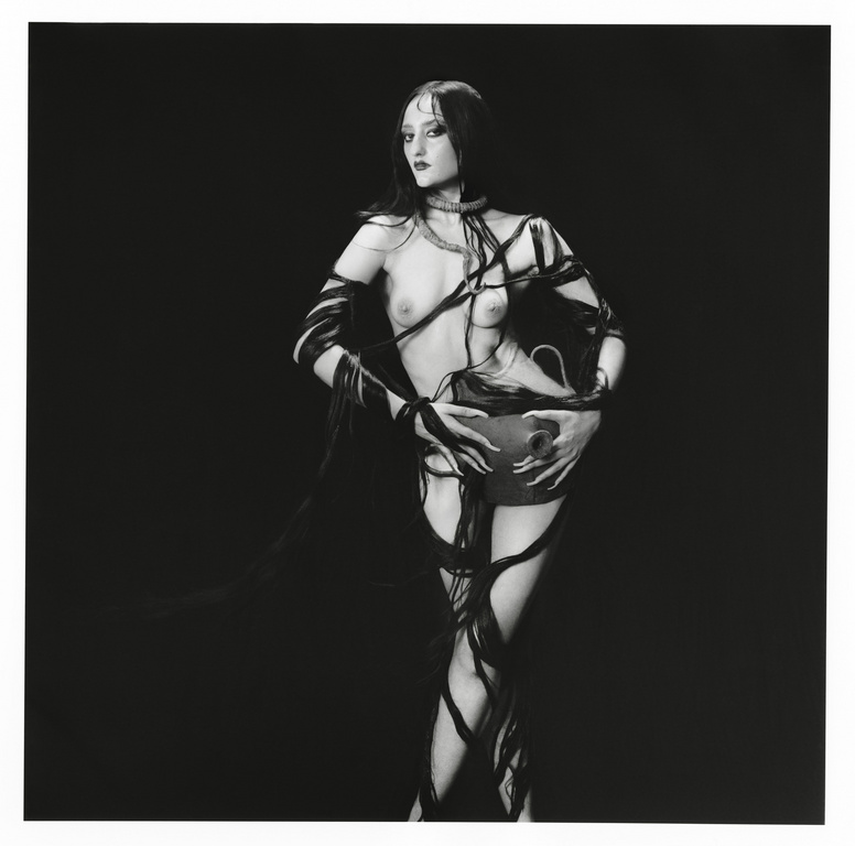 Woman in dark makeup and ribbons holding a dark vase in a black and white photograph, posing in front of a black background.