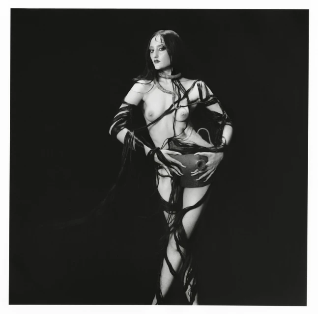 Woman in dark makeup and ribbons holding a dark vase in a black and white photograph, posing in front of a black background.