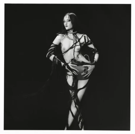 Woman in dark makeup and ribbons holding a dark vase in a black and white photograph, posing in front of a black background.