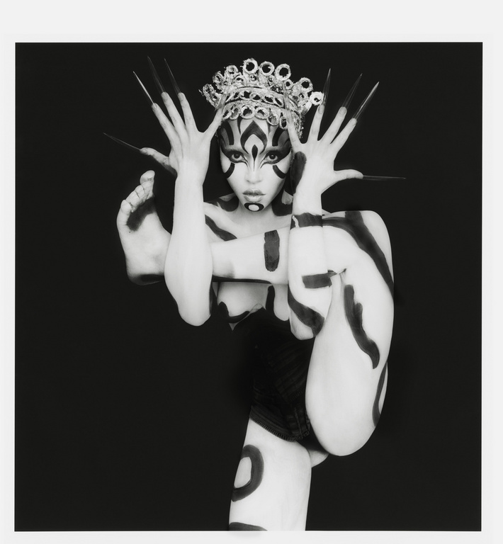 Artistic black-and-white portrait of a person with body paint, a crown, and exaggerated makeup, set against a dark backdrop in a studio environment.