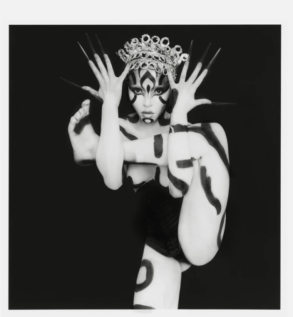 Artistic black-and-white portrait of a person with body paint, a crown, and exaggerated makeup, set against a dark backdrop in a studio environment.