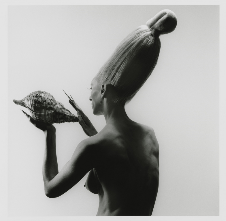 Artistic black and white portrait of a bare-chested adult woman with an elaborate hairstyle, holding a large conch shell.