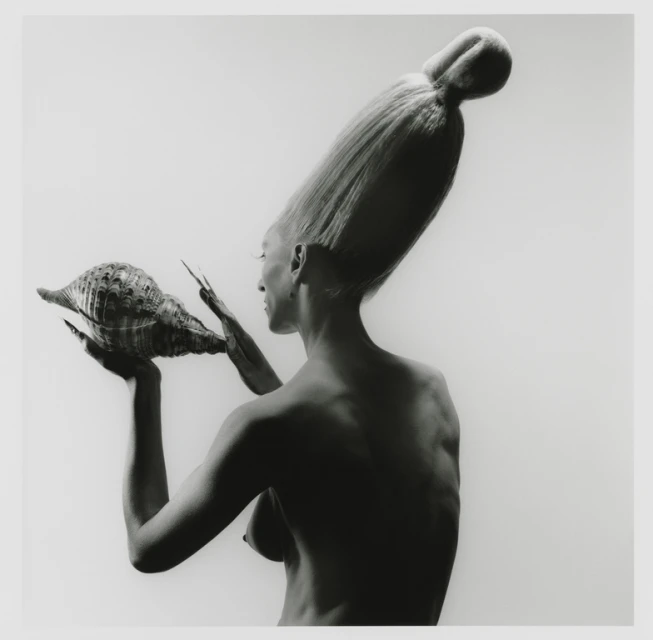 Artistic black and white portrait of a bare-chested adult woman with an elaborate hairstyle, holding a large conch shell.