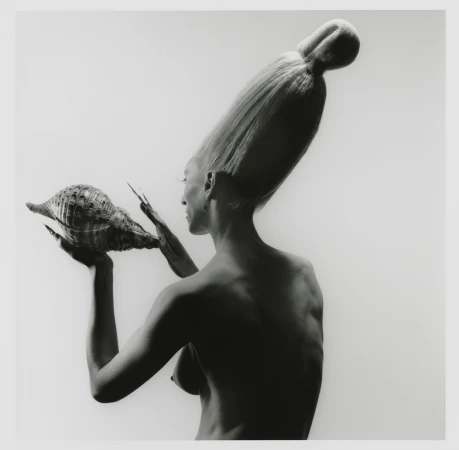 Artistic black and white portrait of a bare-chested adult woman with an elaborate hairstyle, holding a large conch shell.