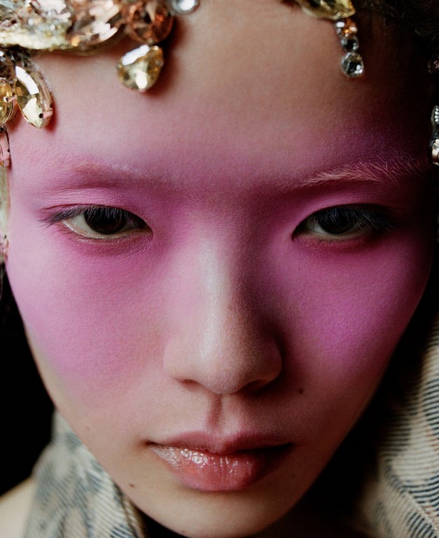 Close-up portrait of an Asian female with bold pink face makeup, decorative jeweled headpiece, and light eye makeup in a studio setting.
