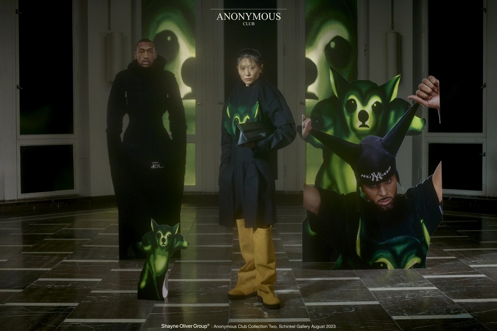 Two models present fashion designs with a printed backdrop and green cutout figures, in an indoor studio showcasing modern apparel.