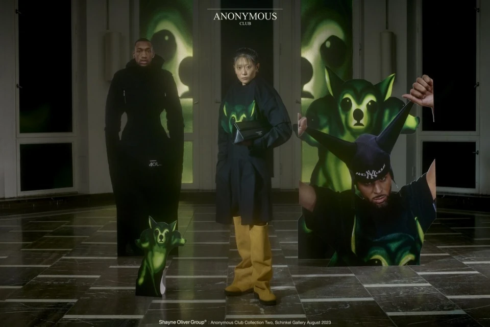 Two models present fashion designs with a printed backdrop and green cutout figures, in an indoor studio showcasing modern apparel.