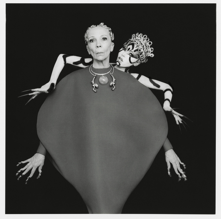 Stylized studio portrait features an older adult female with an elaborately costumed female figure behind her, captured in a black and white aesthetic.