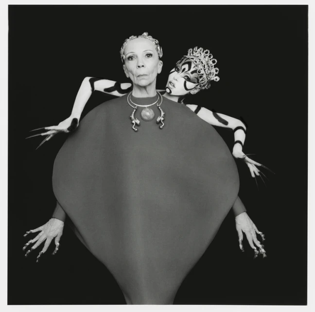 Stylized studio portrait features an older adult female with an elaborately costumed female figure behind her, captured in a black and white aesthetic.