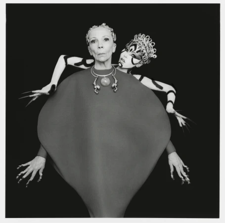 Stylized studio portrait features an older adult female with an elaborately costumed female figure behind her, captured in a black and white aesthetic.