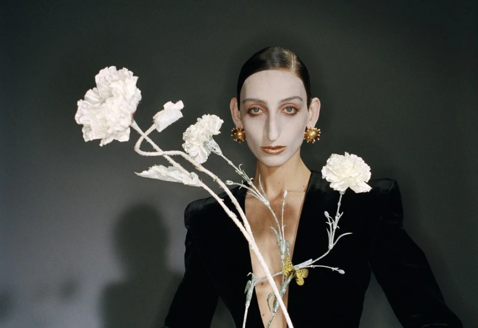 Striking fashion portrait of a person with dramatic makeup and large earrings, holding a white floral arrangement against a dark gray backdrop.
