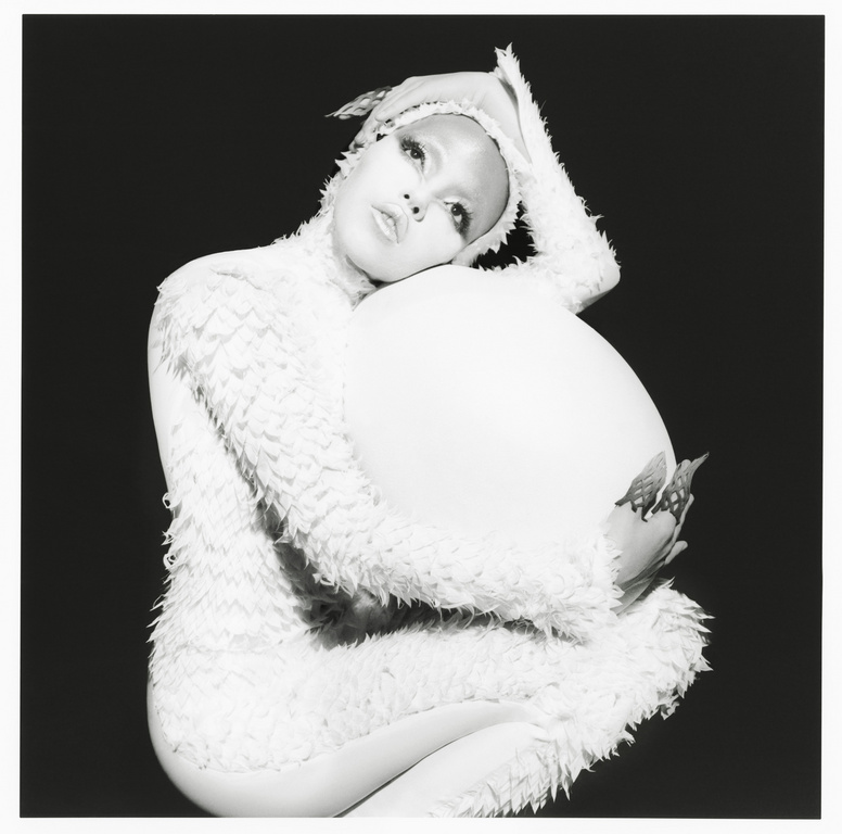 Black and white studio portrait of a person with artistic makeup and a feathered costume, cradling a large white prop.