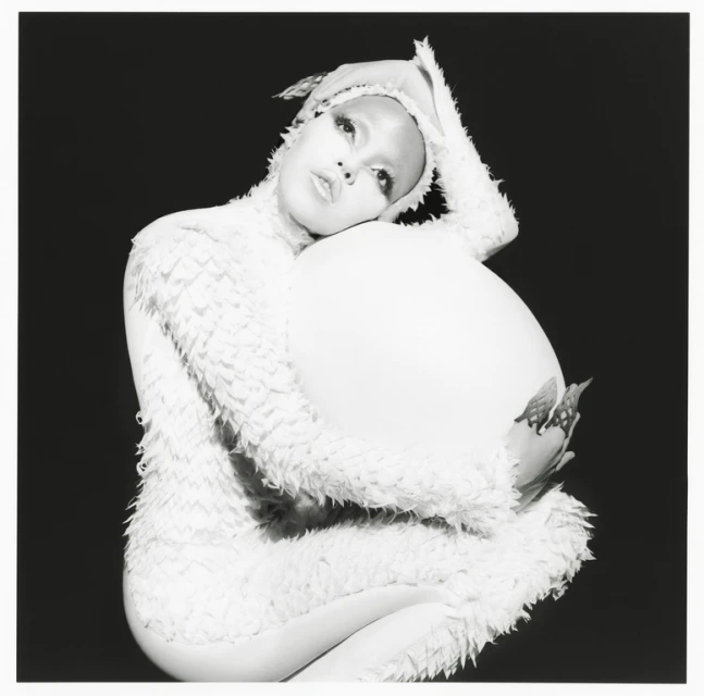 Black and white studio portrait of a person with artistic makeup and a feathered costume, cradling a large white prop.