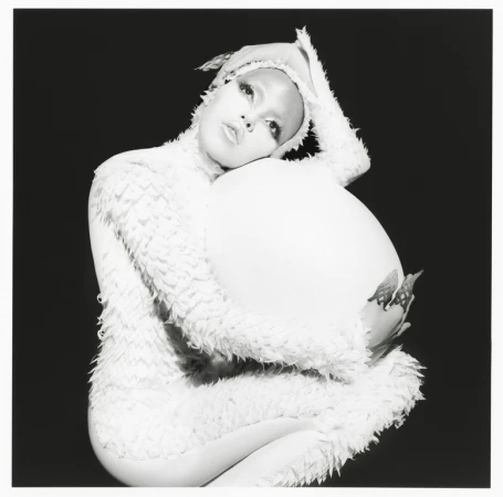 Black and white studio portrait of a person with artistic makeup and a feathered costume, cradling a large white prop.