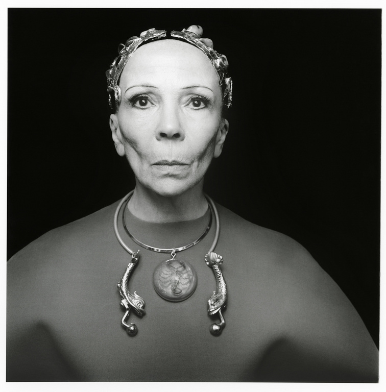 Black and white portrait of an adult woman wearing a metallic headpiece and an ornamental necklace against a black background.