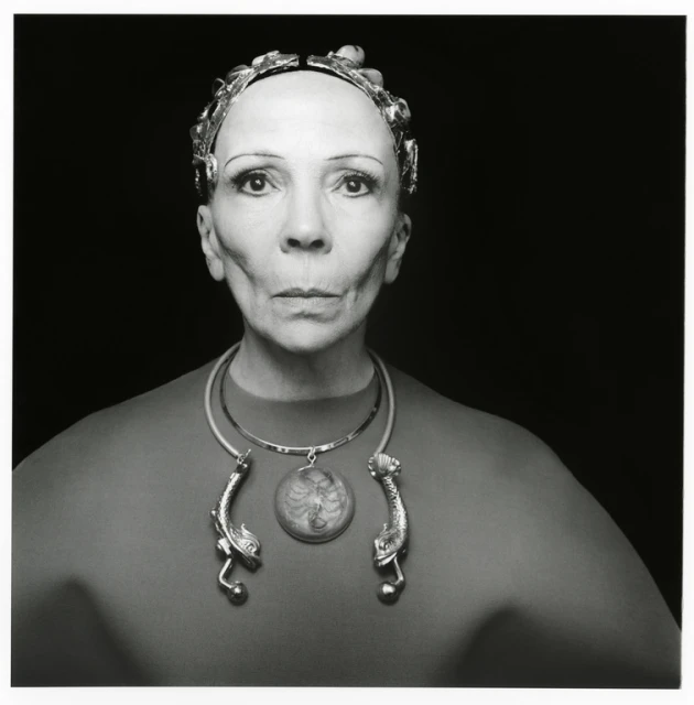 Black and white portrait of an adult woman wearing a metallic headpiece and an ornamental necklace against a black background.