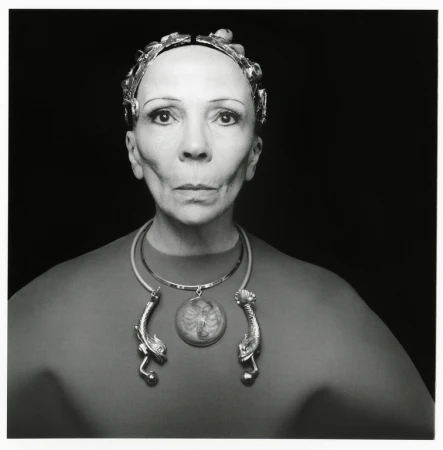 Black and white portrait of an adult woman wearing a metallic headpiece and an ornamental necklace against a black background.