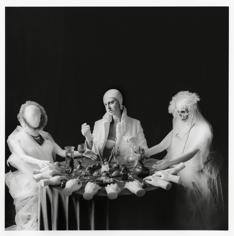 Three posed figures in white around a table covered with mannequin hands, glassware, and food against a dark studio backdrop in black and white.