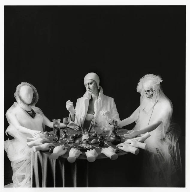Three posed figures in white around a table covered with mannequin hands, glassware, and food against a dark studio backdrop in black and white.