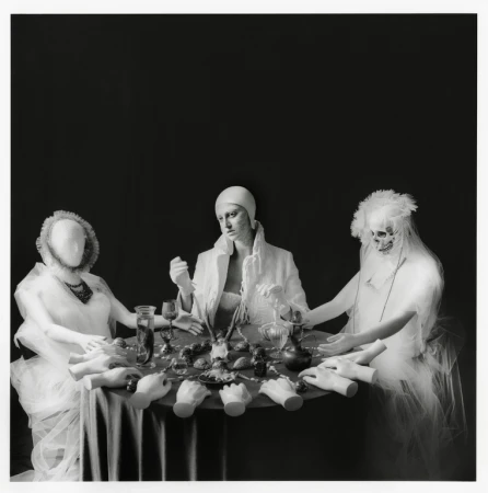 Three posed figures in white around a table covered with mannequin hands, glassware, and food against a dark studio backdrop in black and white.