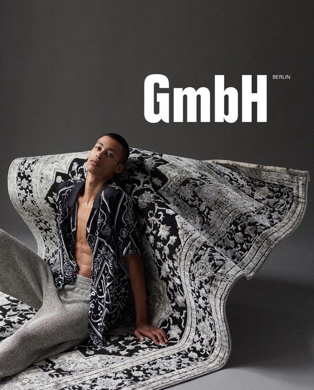 An adult male model poses indoors, wearing a patterned button-down shirt and pants, lying on a patterned rug, against a grey backdrop.