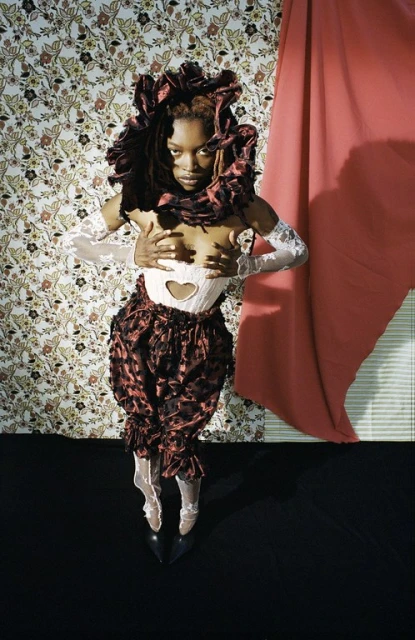 Eye-level view of an adult fashion model in a ruffled collar and bloomers posing in front of floral wallpaper and a red curtain.