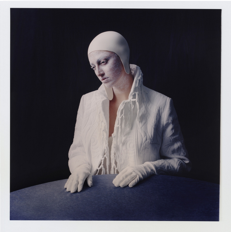 Woman with pale skin, wearing white head covering, white gloves, and a textured white jacket, poses in front of a dark background.