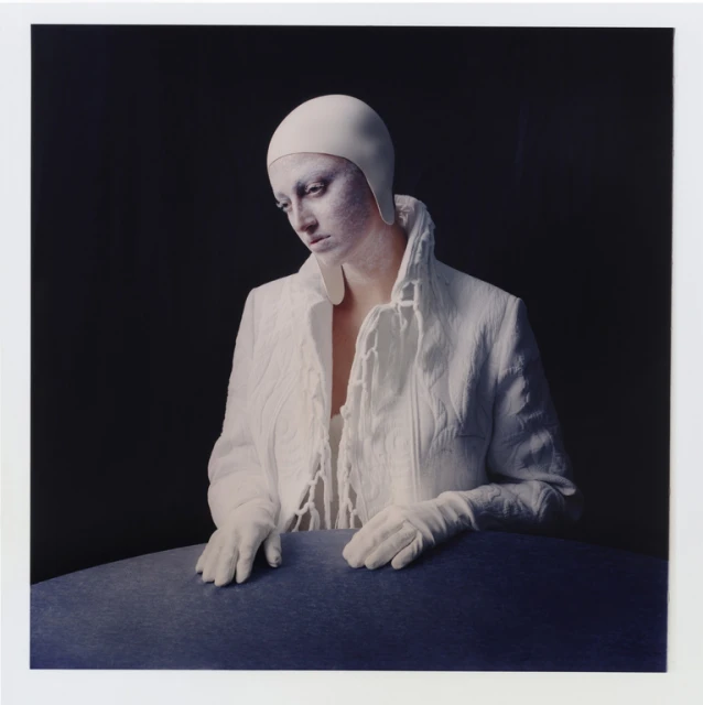 Woman with pale skin, wearing white head covering, white gloves, and a textured white jacket, poses in front of a dark background.