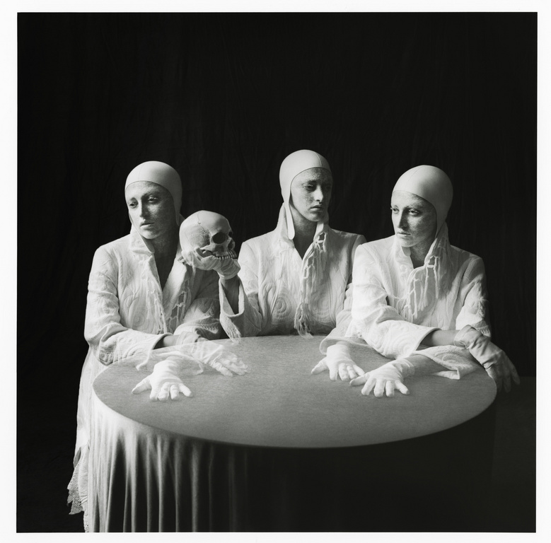 Three pale women in white surgical gowns and caps pose at a round table with a skull in a darkly lit, high-contrast fine art photo.