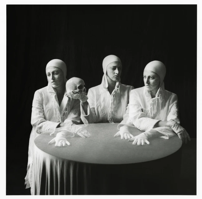 Three pale women in white surgical gowns and caps pose at a round table with a skull in a darkly lit, high-contrast fine art photo.