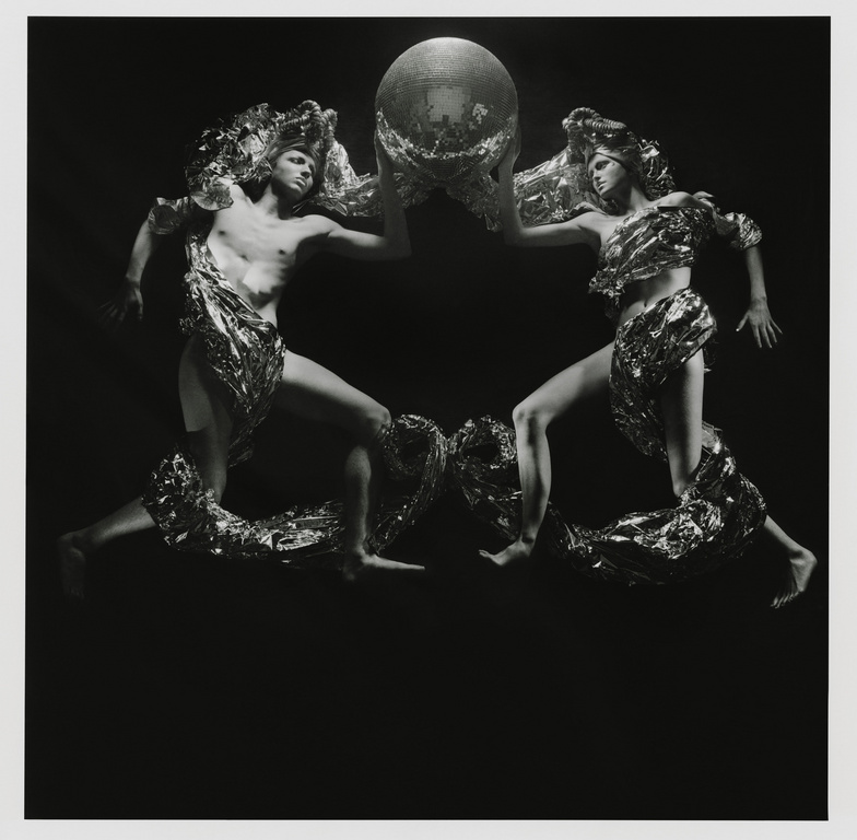 Two figures draped in metallic fabric hold a mirrored disco ball in a high-contrast black and white studio photography composition.