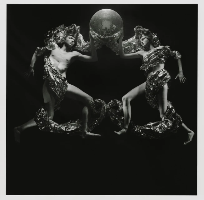 Two figures draped in metallic fabric hold a mirrored disco ball in a high-contrast black and white studio photography composition.