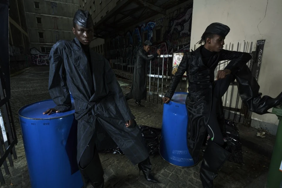 Three models in dark jackets and caps pose next to blue barrels against a graffiti-covered wall. Urban fashion in dim, gritty, outdoor scene.