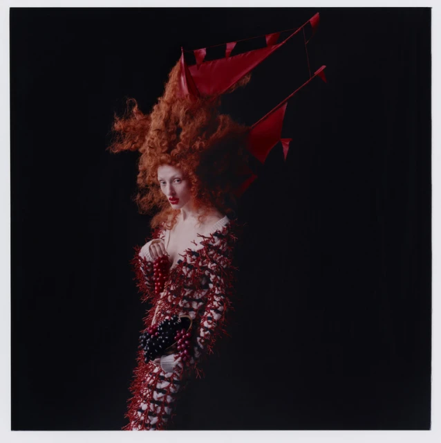 Woman with red hair and avant-garde fashion poses in a studio against a black backdrop with dramatic lighting in a square composition.