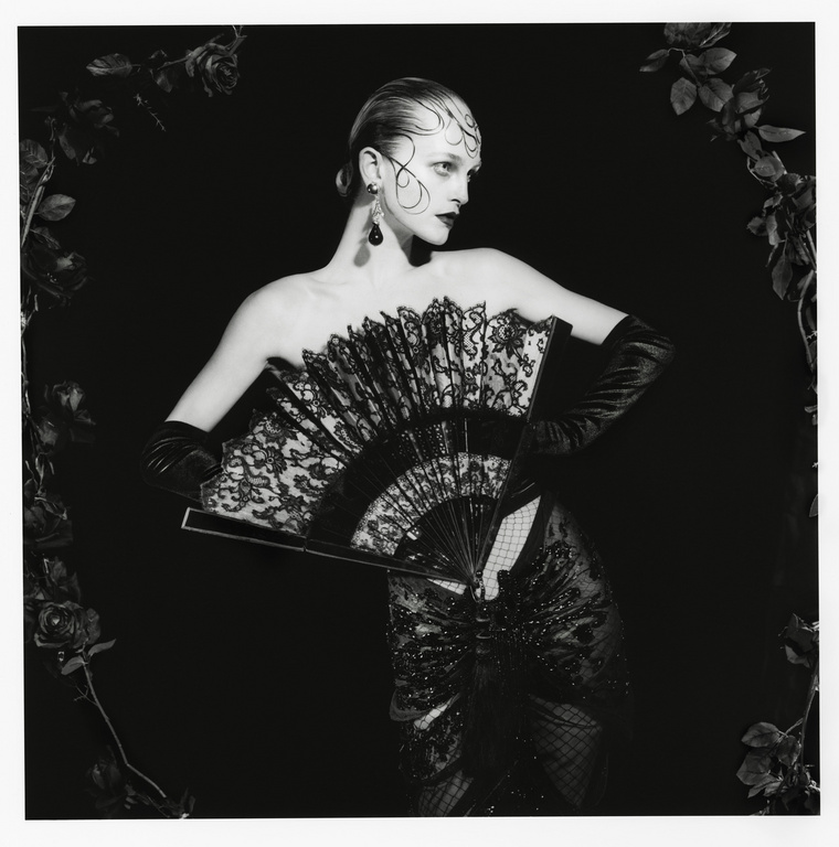 Woman in strapless black gown and gloves holds a fan, framed by dark roses against a black backdrop for a dramatic fashion portrait.