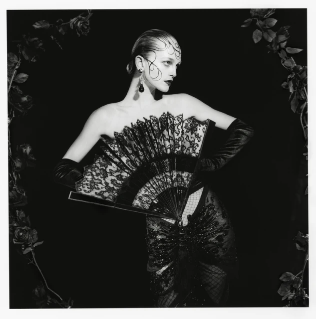 Woman in strapless black gown and gloves holds a fan, framed by dark roses against a black backdrop for a dramatic fashion portrait.