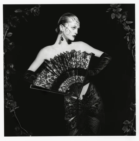Woman in strapless black gown and gloves holds a fan, framed by dark roses against a black backdrop for a dramatic fashion portrait.