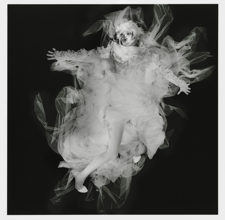 Artistic fashion portrait featuring a person in elaborate white tulle costume against a stark black background, creating a dramatic visual.