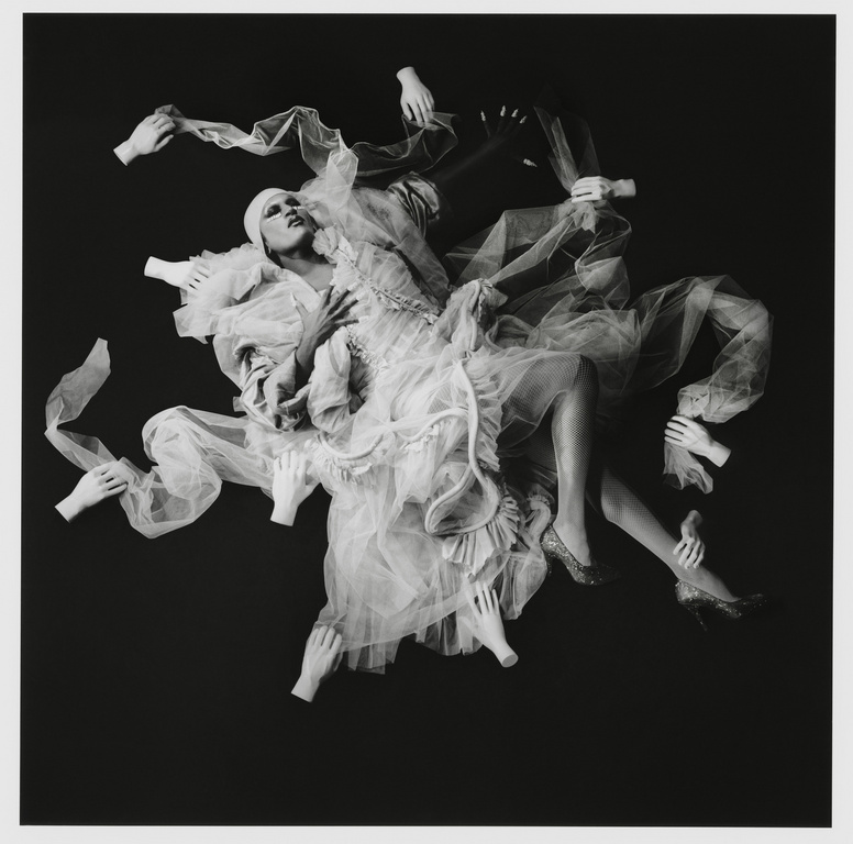Surreal black and white studio portrait of a person in frilly white dress surrounded by floating disembodied hands against a dark background.