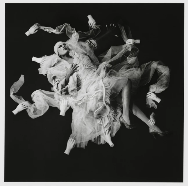 Surreal black and white studio portrait of a person in frilly white dress surrounded by floating disembodied hands against a dark background.