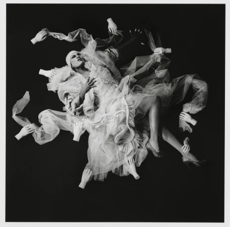 Surreal black and white studio portrait of a person in frilly white dress surrounded by floating disembodied hands against a dark background.